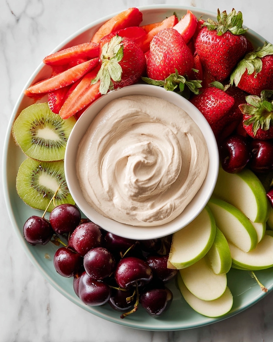 A white round plate with a smaller white bowl in the center filled with creamy, light beige dip smooth with soft swirls. Around the bowl are fresh fruit pieces arranged in sections: bright red strawberries with green leaves, bright green kiwi slices with their seeds showing, deep red cherries, dark red and purple grapes with a slight shine, red plum wedges, and thin green apple slices. The plate sits on a white marbled surface. photo taken with an iphone --ar 4:5 --v 7