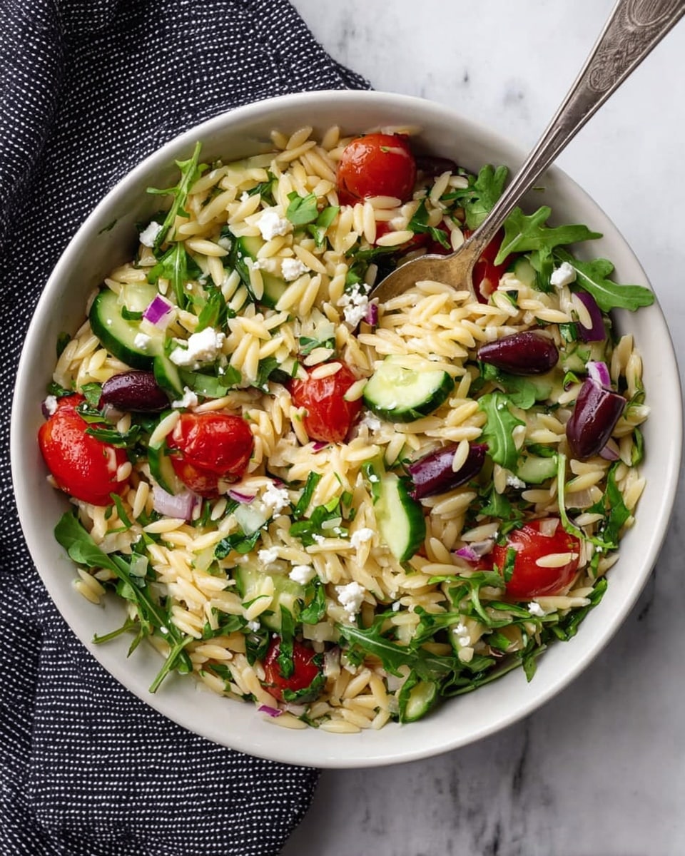A white bowl filled with a colorful orzo salad shows three main layers: a base of small, light yellow orzo pasta with a smooth texture, mixed evenly with bright green arugula leaves scattered throughout as the second layer. On top, there are chunky, fresh pieces of red cherry tomatoes, dark purple olives, crisp green cucumber slices, and small white feta cheese crumbles adding variety in color and texture. A silver spoon rests inside the bowl, partially covered by the salad. The bowl sits on a black and white checkered cloth over a white marbled surface. Photo taken with an iphone --ar 4:5 --v 7