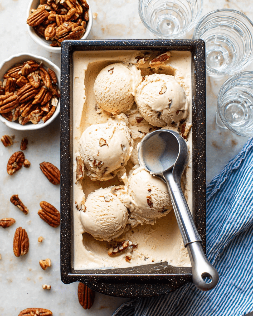 A close-up top view of a rectangular black-speckled container filled with light beige ice cream with visible pecan pieces mixed in, topped with five scoops of the same ice cream arranged unevenly; a silver ice cream scoop rests inside the container on the right side. Scattered around the container are pecan halves, with a small white bowl filled with pecans placed on the left side. Two clear glass dessert cups sit at the top right corner on a white marbled texture surface, along with a white cloth with blue stripes folded underneath the container on the bottom right side. photo taken with an iphone --ar 4:5 --v 7