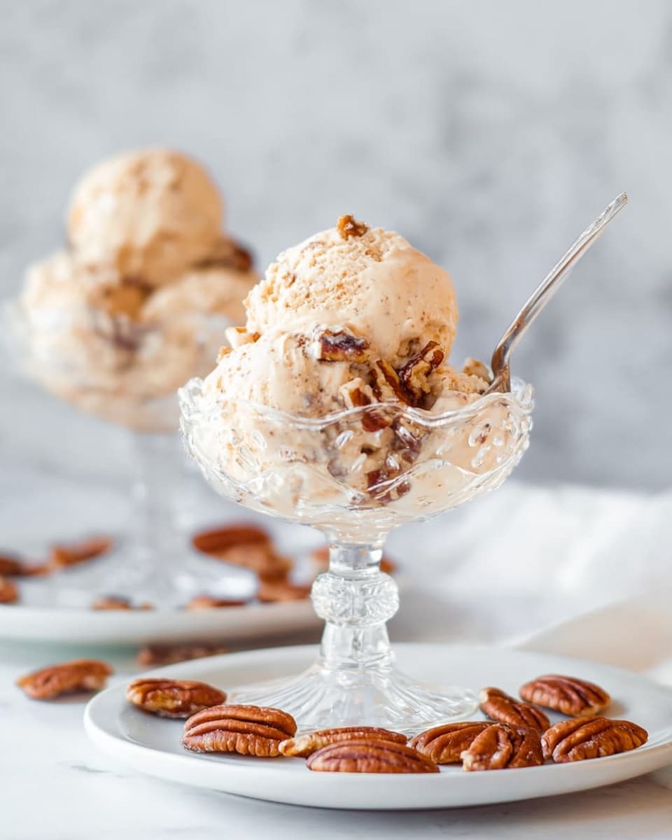The image shows two clear glass dessert dishes filled with two scoops each of light beige ice cream with visible pecan pieces mixed inside. The dishes have a scalloped edge and a stem base, placed on a white plate scattered with pecan halves. One dish is in the front and sharply focused, with a silver spoon inserted in the ice cream, while the other dish is blurred in the background. The setting is on a white marbled texture with a soft, out-of-focus background. photo taken with an iphone --ar 4:5 --v 7
