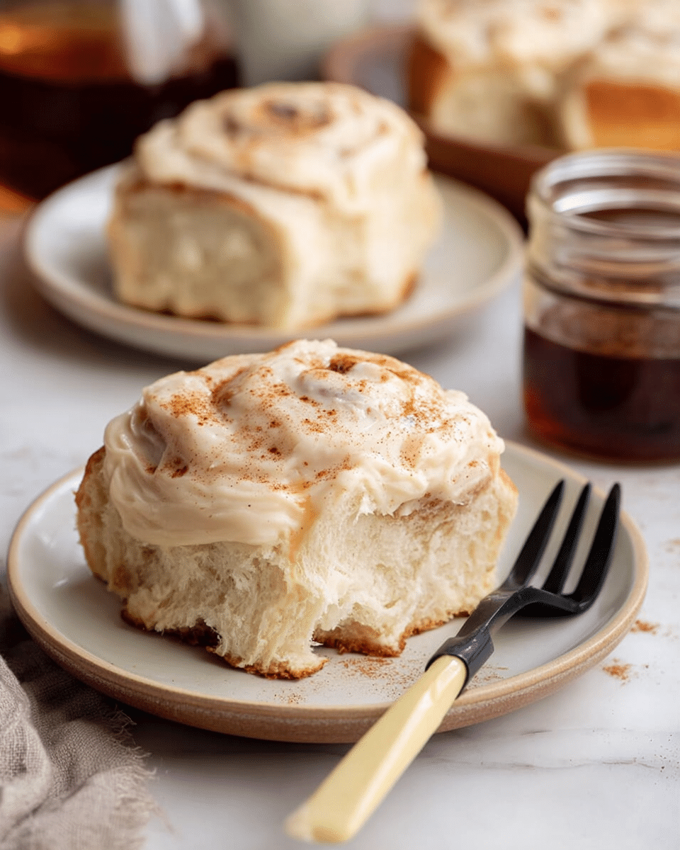 A close-up of a soft, fluffy cinnamon roll placed on a small white plate, showing two visible layers of light golden-brown dough with a thick, creamy layer of pale beige frosting generously spread on top, sprinkled lightly with cinnamon. The cinnamon roll has a slightly torn edge exposing its airy texture. In the background, there are two more cinnamon rolls on white plates and a glass jar with cinnamon powder. A black fork with a pale yellow handle rests on the edge of the plate, and a glass of dark syrup is partially visible on the left. All items sit on a white marbled surface. Photo taken with an iphone --ar 4:5 --v 7