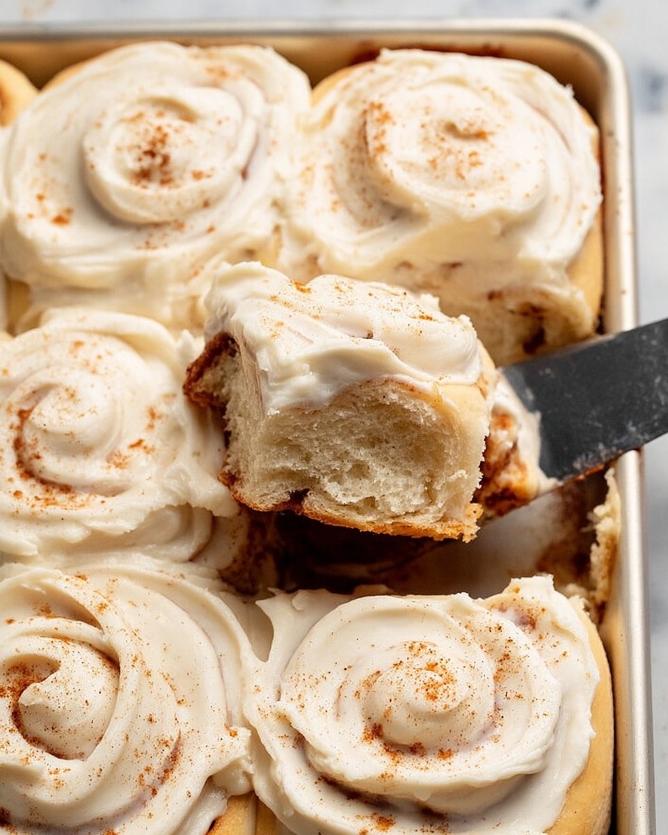A close-up view of a tray filled with soft, golden brown cinnamon rolls arranged in two rows. Each roll is generously covered with a thick, creamy white frosting that has smooth swirls and a slightly glossy texture. The frosting has a light sprinkle of cinnamon powder on top, adding a touch of warm brown color. One cinnamon roll is being lifted with a shiny silver spatula, revealing a fluffy inner dough with a speckled cinnamon filling. The tray rests on a white marbled surface. photo taken with an iphone --ar 4:5 --v 7