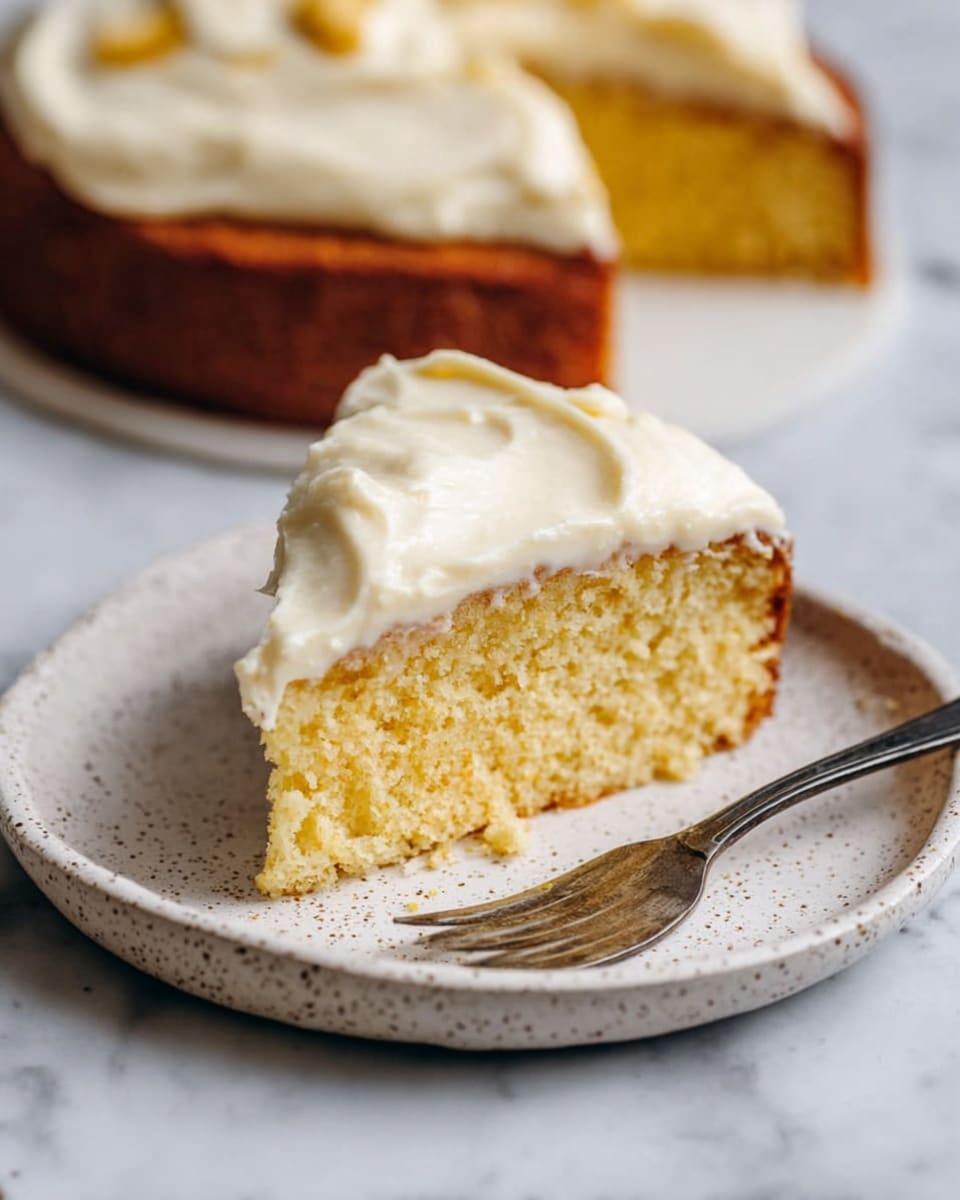 A single slice of yellow cake with a slightly crumbly texture sits on a white speckled plate, topped with a thick, creamy layer of pale off-white frosting that looks soft and smooth. A silver fork rests on the plate next to the slice. In the background, the remaining cake is visible with a golden-brown crust and the same creamy topping, all placed on a white marbled surface. Photo taken with an iphone --ar 4:5 --v 7
