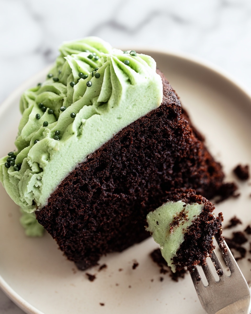 A close-up of a single slice of dark chocolate cake with a rich, moist texture, topped with a thick layer of light green frosting that is piped with decorative swirls and small round sprinkles scattered on top. The cake slice sits on a plain white plate with some cake crumbs around it. To the right of the cake slice, there is a fork with bits of chocolate cake and green frosting on its tines, resting on the plate. The background has a white marbled texture. photo taken with an iphone --ar 4:5 --v 7