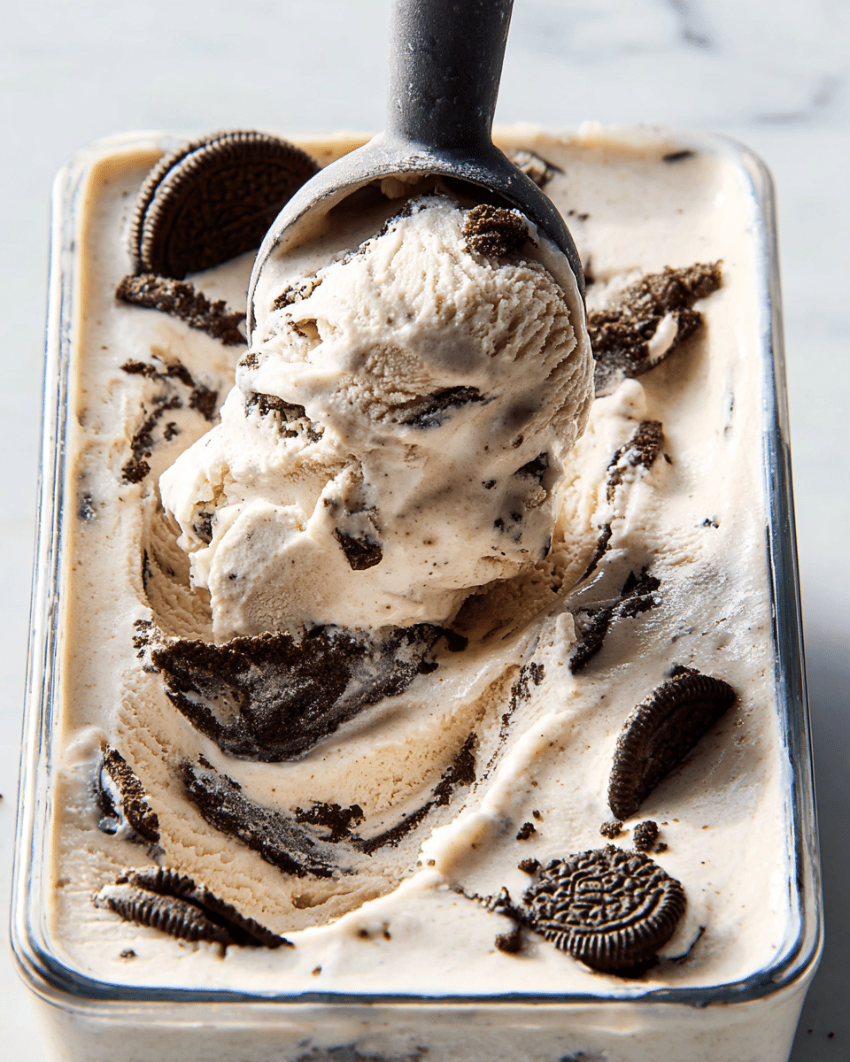 The image shows a glass dessert cup filled with creamy cookies and cream ice cream, which is light beige with small dark cookie bits mixed inside. On top, there is a thick layer of white whipped cream that looks soft and fluffy. Both the ice cream and whipped cream have several pieces of broken chocolate sandwich cookies scattered over them, adding a rough texture and dark brown color contrast. A silver spoon is scooping up some of the ice cream from the right side of the cup. The dessert cup sits on a white marbled surface with a blurry greyish background. photo taken with an iphone --ar 4:5 --v 7