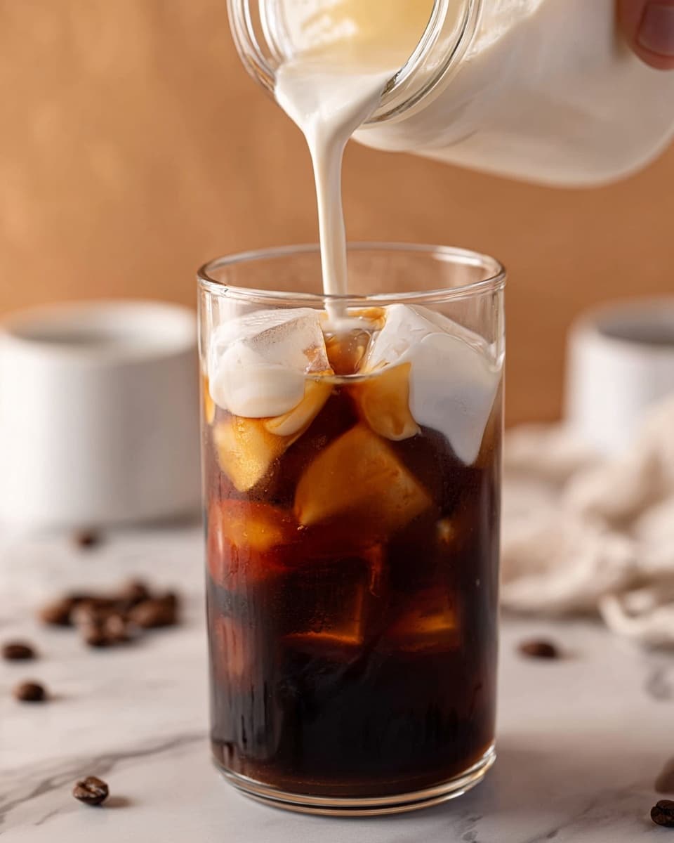A clear glass filled with dark brown iced coffee and large translucent ice cubes, topped with thick, creamy white milk being poured from above. The drink shows two main layers: the dark bottom coffee with ice and the soft white milk foam layer forming at the top. The background has a warm, soft tone with scattered coffee beans and white cups slightly blurred. A woman's hand is holding the container from which the milk is poured. The surface is a white marbled texture. photo taken with an iphone --ar 4:5 --v 7
