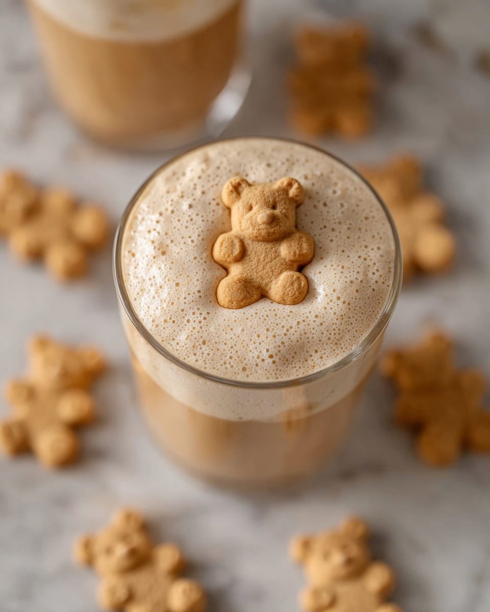 The image shows a glass filled with a creamy, light brown whipped coffee drink with a foamy texture on top. In the center of the foam sits a small pale beige teddy bear-shaped cookie. In the background, another similar glass is partially visible, also topped with the same foam and a teddy bear cookie. Around the base of the glasses, multiple teddy bear cookies lie scattered on a white marbled surface. photo taken with an iphone --ar 4:5 --v 7