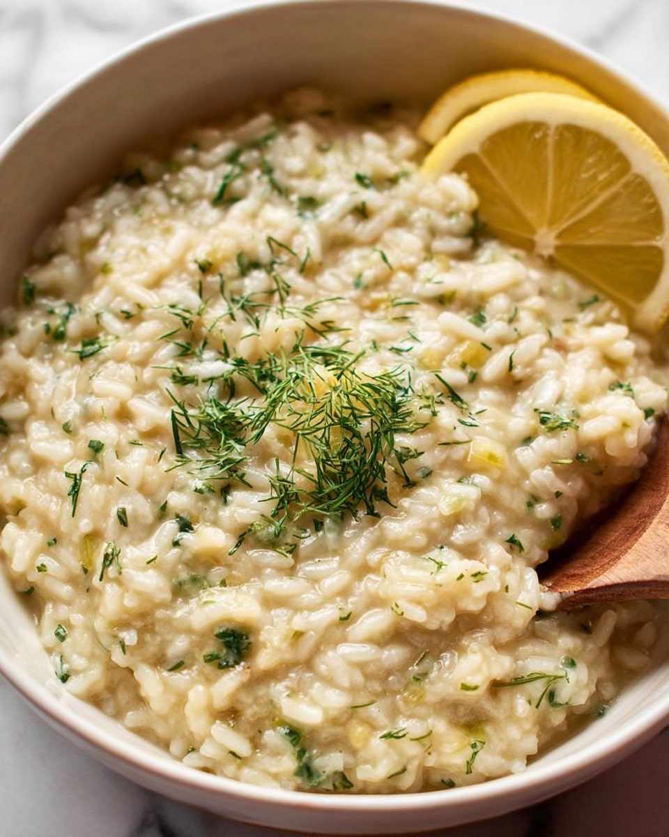 The image shows a close-up of a dish with two main layers. The bottom layer is creamy white risotto mixed with finely chopped green herbs, giving it a soft and smooth texture with small grain shapes. On top lies a piece of cooked fish with a crisp, brown, and textured skin, revealing tender pinkish flesh underneath. The fish has a sprinkle of coarse salt on the crispy skin. To the right, there is a white porcelain spoon partly submerged in the risotto, lifting some of it. A slice of bright yellow lemon is visible in the background, placed near the fish. The entire dish is served on a white plate on a white marbled surface. Photo taken with an iphone --ar 4:5 --v 7