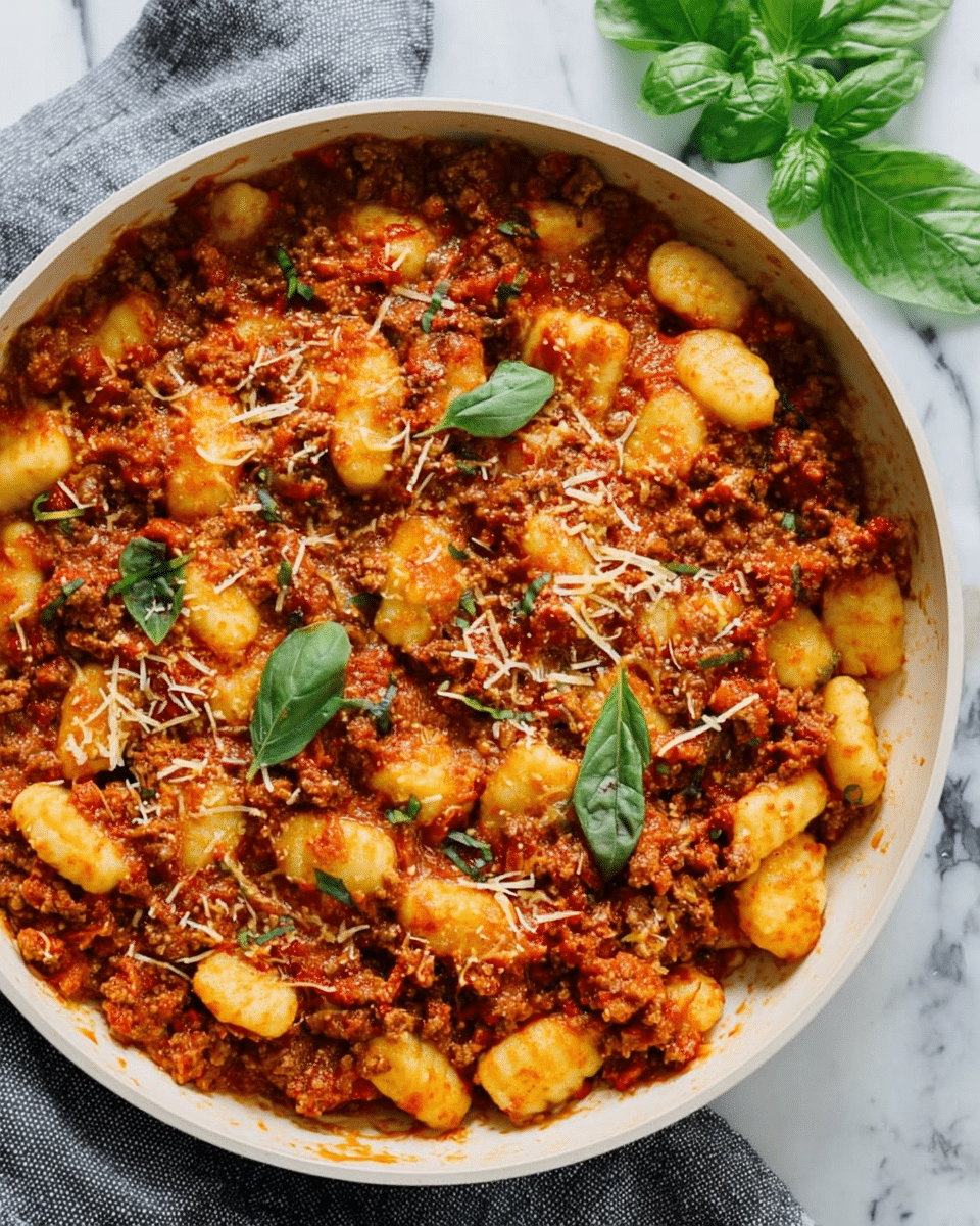 A white pan filled with gnocchi covered in a thick, chunky red meat sauce. The gnocchi are pale yellow with a soft, slightly ridged texture and are mixed evenly in the sauce, which has a rich reddish-brown color with visible bits of ground meat and small green herb pieces scattered throughout. On top, there are fine shreds of light yellow cheese sprinkled over the dish. In the upper right corner, there are fresh green basil leaves placed as a garnish. The pan is placed on a white marbled surface with a gray cloth partially visible at the bottom left corner. photo taken with an iphone --ar 4:5 --v 7