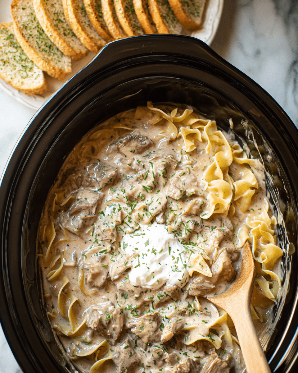 This image shows a black slow cooker filled with a creamy beef stroganoff mixture. The dish has three main layers: wide egg noodles that are light yellow and slightly ruffled, tender chunks of brown beef mixed evenly, and a thick beige sauce coated over everything giving a smooth texture. On the top center, there is a dollop of white sour cream sprinkled with green herbs. A wooden spoon is partially dipped into the mix on the right side. In the background, partially visible, there is a white plate stacked with slices of toasted bread with green herb seasoning. The entire scene is set on a white marbled surface. photo taken with an iphone --ar 4:5 --v 7