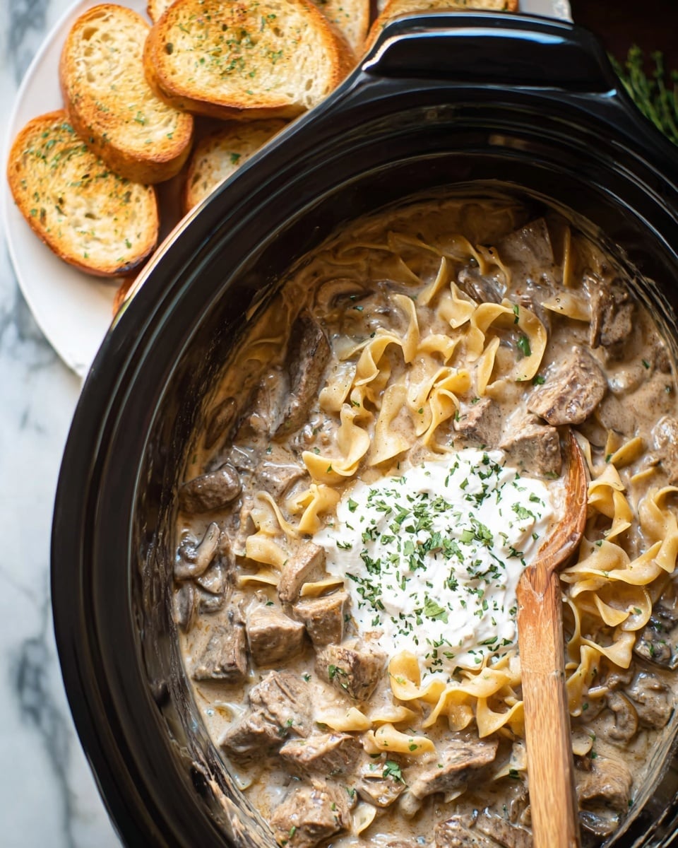 A close-up top view of a slow cooker filled with creamy beef stroganoff, showing three main layers: a base layer of tender chunks of brown beef mixed with sliced mushrooms in a thick beige sauce, a middle layer of soft egg noodles coated in the same sauce, and a dollop of white sour cream sprinkled with green parsley in the center. A wooden spoon with sauce clings to it rests inside the pot on the right side. In the background, there is a white plate holding several slices of toasted garlic bread. The whole scene is set on a white marbled surface. Photo taken with an iphone --ar 4:5 --v 7