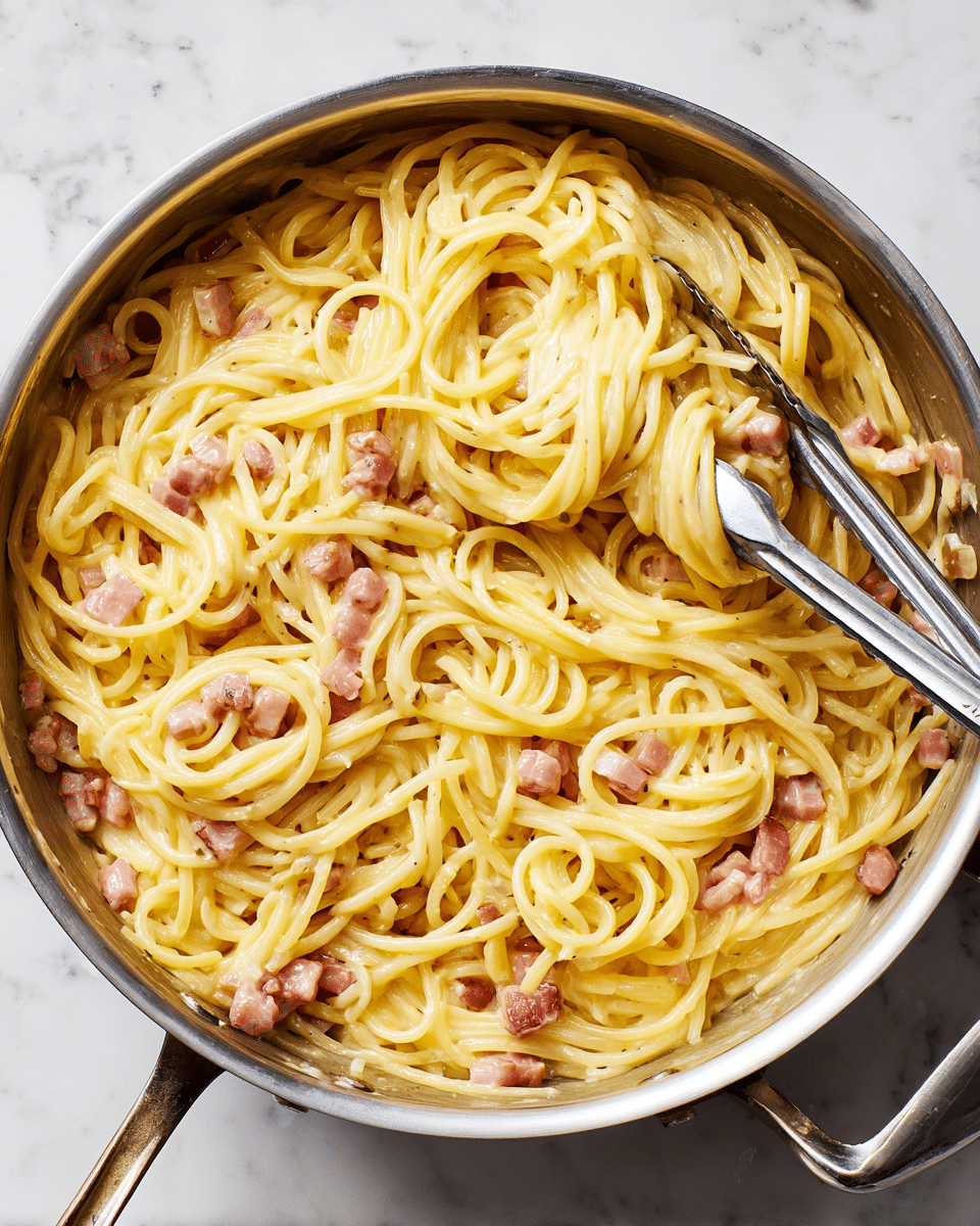 A large stainless steel pan filled with creamy yellow spaghetti mixed with small chunks of pinkish cooked bacon. The pasta is shiny and coated with a smooth, thick sauce that lightly clings to each strand. The bacon pieces are scattered evenly throughout the dish. A pair of silver metal tongs rests inside the pan, with some pasta twisted around the tongs. The pan is placed on a white marbled texture surface. photo taken with an iphone --ar 4:5 --v 7