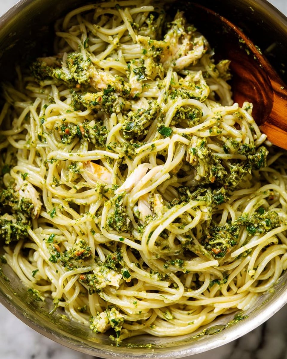 A close-up view of a large pile of cooked thin noodles mixed with a green pesto-like sauce that has a slightly chunky texture, with visible bits of herbs and small pieces of a pale ingredient, possibly chicken or tofu, spread evenly throughout the noodles. The noodles are light yellowish-white, and the green sauce adds a fresh and textured contrast. The dish is shown inside a metal bowl with a wooden spoon partially visible on one side, resting among the noodles. The background is a white marbled texture. photo taken with an iphone --ar 4:5 --v 7