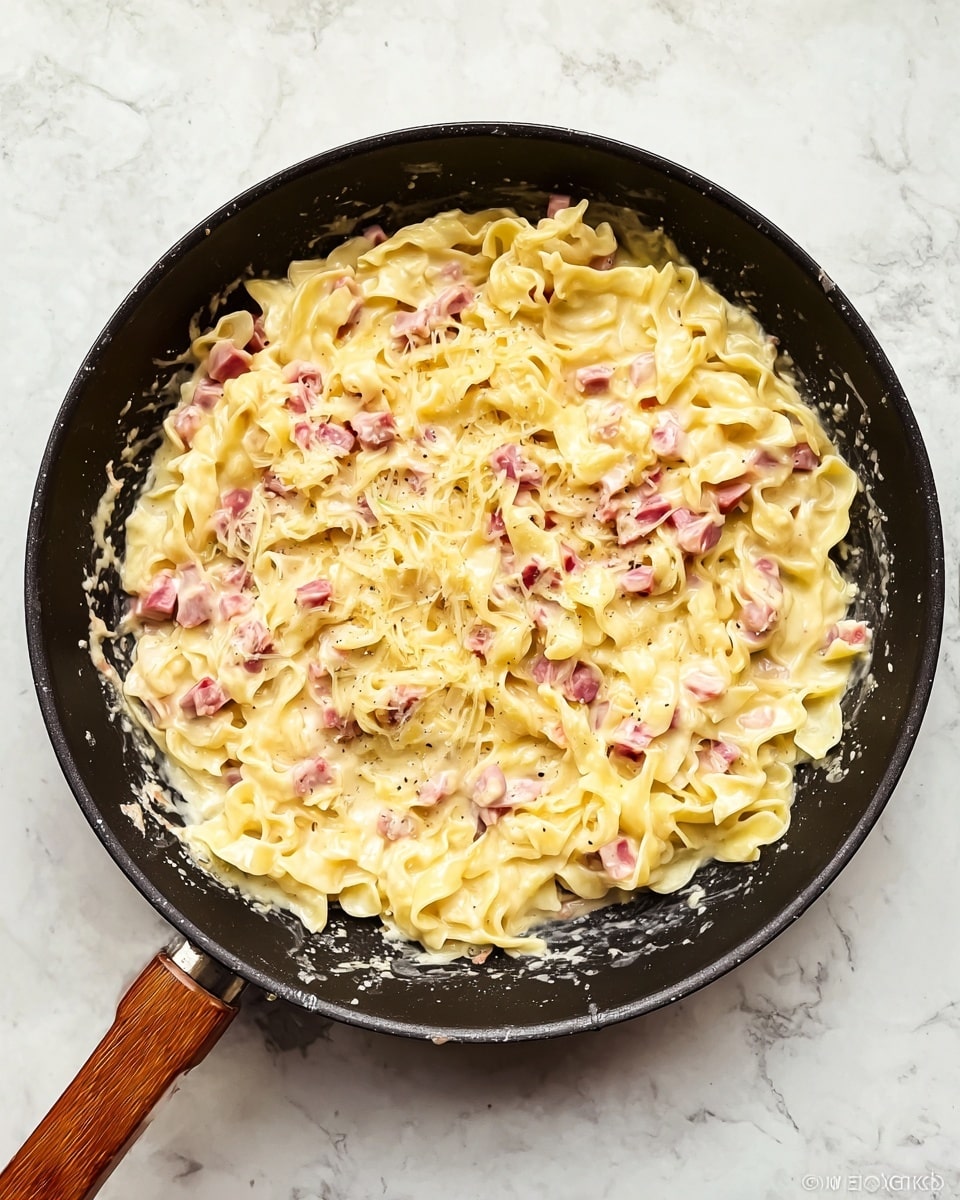 The image shows a black pan with a wooden handle filled with creamy pasta. The pasta is made of flat, slightly curled yellow noodles mixed with small chunks of pink ham, all coated in a thick, pale yellow cheese sauce. The creamy sauce covers the pasta evenly, giving it a smooth texture with a few slightly browned edges. The pan is placed on a white marbled surface, and the image is taken from above, capturing the whole pan and some splashes of sauce on its edge. photo taken with an iphone --ar 4:5 --v 7