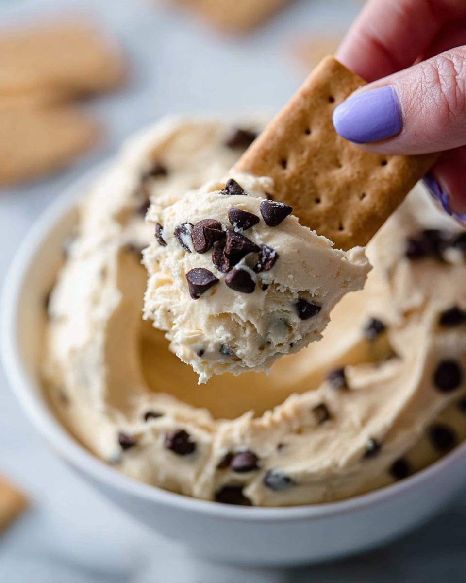A close-up view of a white bowl filled with thick, creamy cookie dough that is pale beige in color with scattered dark chocolate chips. A rectangular, textured cracker of light brown color is held by a woman's hand with purple painted nails, scooping up a generous scoop of the dough studded with shiny chocolate chips. The background shows more creamy dough inside the bowl with a soft focus white marbled surface. photo taken with an iphone --ar 4:5 --v 7